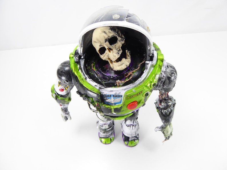 May include: A modified Buzz Lightyear action figure with a skull inside the helmet. The figure is primarily green, black, and white, with the words "Space Ranger" and "Lightyear" visible. The figure has a distressed, zombie-like appearance.