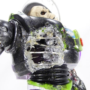 May include: A modified action figure of a space ranger, with a skeletal head visible inside the helmet. The figure's armor is black, green, and white, with visible damage and exposed internal components. The word "LASER" is visible.