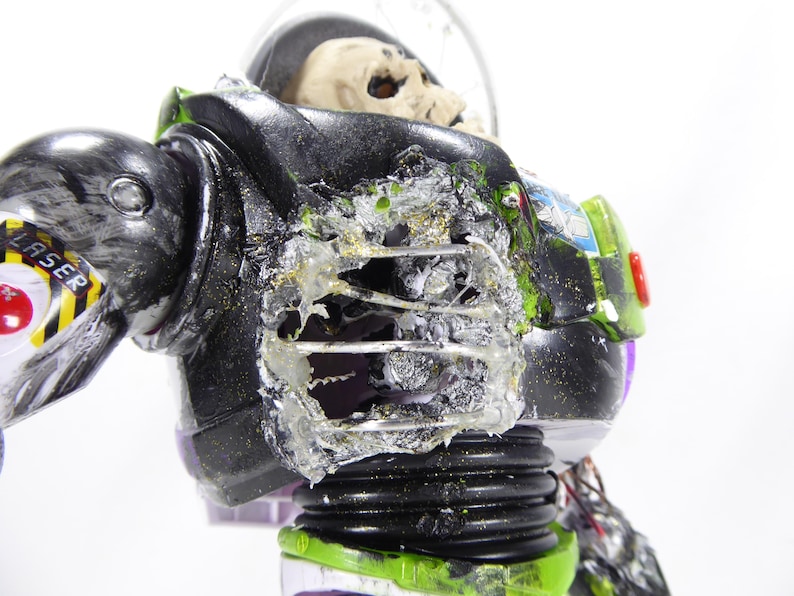 May include: A modified action figure of a space ranger, with a skeletal head visible inside the helmet. The figure's armor is black, green, and white, with visible damage and exposed internal components. The word "LASER" is visible.