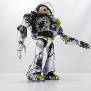 May include: A modified Buzz Lightyear action figure with a skeletal head inside the helmet. The toy has a black, white, green, and purple color scheme, with exposed wiring and battle damage details. The figure is posed against a white background.
