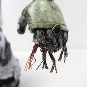 May include: A close-up of a grotesque, weathered sculpture of a hand. The hand is painted in shades of green and black, with exposed red and copper-colored wires extending from the wrist. The sculpture has a distressed, aged appearance.