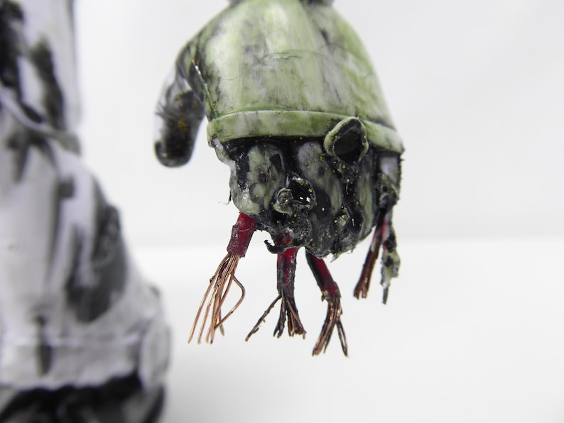 May include: A close-up of a grotesque, weathered sculpture of a hand. The hand is painted in shades of green and black, with exposed red and copper-colored wires extending from the wrist. The sculpture has a distressed, aged appearance.