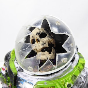 May include: A novelty figurine of an astronaut with a cracked helmet revealing a skull. The astronaut suit is green, black, and silver, with the words "SPACE RANGER" visible. The helmet is clear plastic.