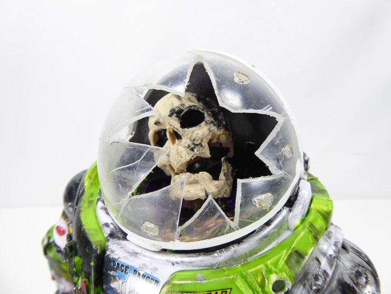 May include: A novelty figurine of an astronaut with a cracked helmet revealing a skull. The astronaut suit is green, black, and silver, with the words "SPACE RANGER" visible. The helmet is clear plastic.
