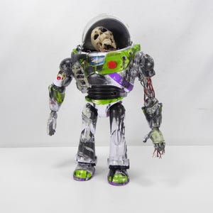 May include: A modified Buzz Lightyear action figure with a skeletal head inside the helmet. The toy is painted in a zombie-like style, with exposed wires and decaying details. The figure's colors include green, white, black, and purple.