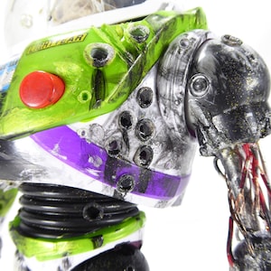 May include: Close-up of a distressed Buzz Lightyear action figure. The toy features a green and white spacesuit with a red button, purple accents, and visible damage. The arm is partially detached, revealing wires.