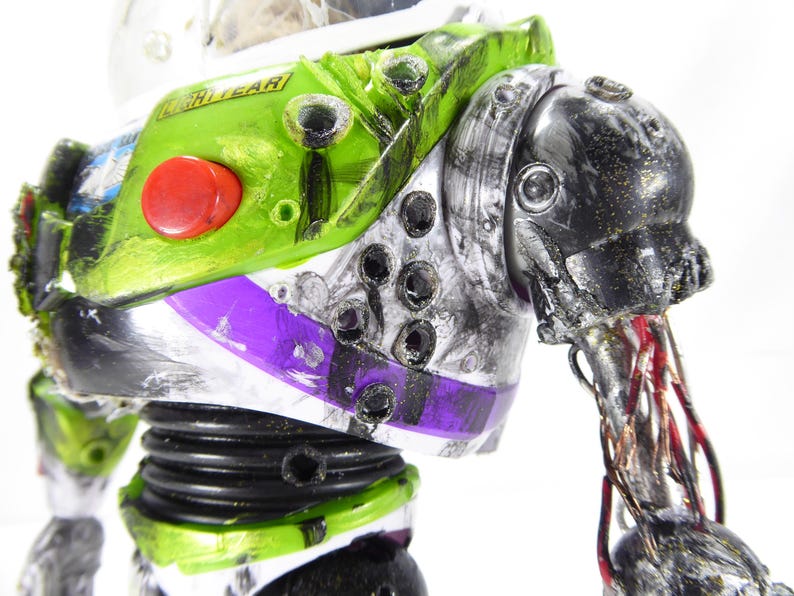 May include: Close-up of a distressed Buzz Lightyear action figure. The toy features a green and white spacesuit with a red button, purple accents, and visible damage. The arm is partially detached, revealing wires.