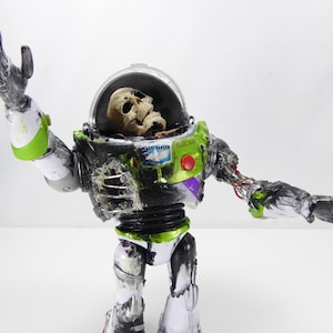 May include: A modified Buzz Lightyear action figure with a skull inside the helmet. The toy is painted in green, white, and black with visible damage and exposed wiring. The figure's arm is outstretched.