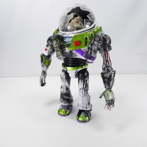 May include: A distressed Buzz Lightyear action figure with a cracked helmet and exposed wiring. The toy features green, white, purple, and black accents, with a red button on the chest. The figure stands on a white surface.