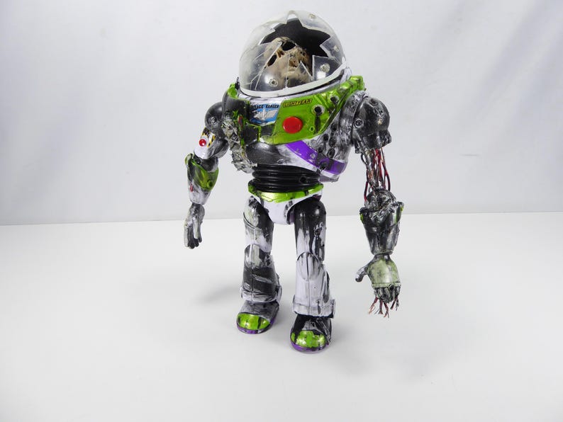 May include: A distressed Buzz Lightyear action figure with a cracked helmet and exposed wiring. The toy features green, white, purple, and black accents, with a red button on the chest. The figure stands on a white surface.