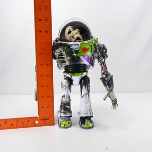 May include: A zombie-themed Buzz Lightyear action figure, approximately 10 inches tall, with a clear helmet revealing a skull. The figure features green, black, and purple accents, with exposed skeletal and decaying details. The figure's right arm is partially detached.
