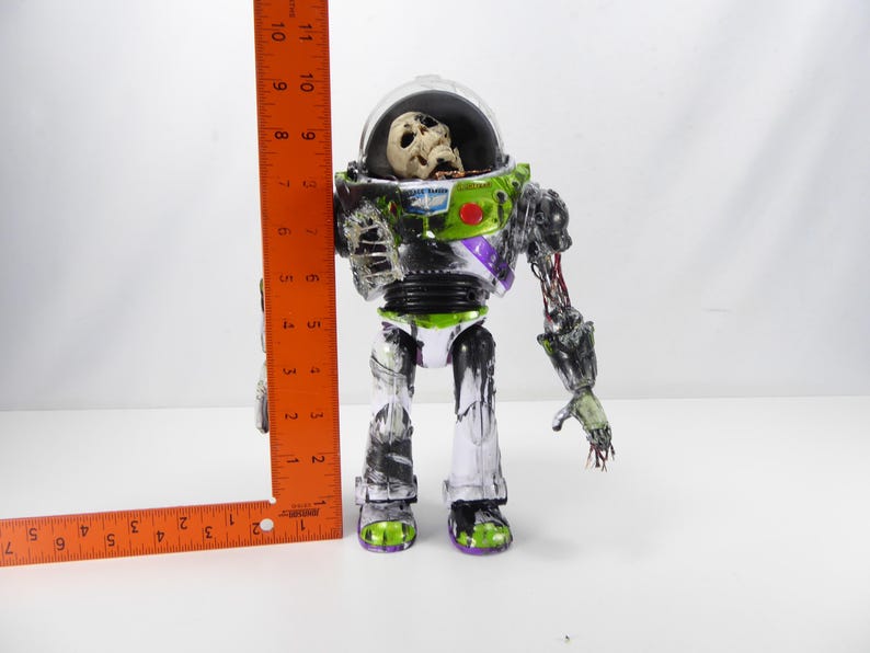 May include: A zombie-themed Buzz Lightyear action figure, approximately 10 inches tall, with a clear helmet revealing a skull. The figure features green, black, and purple accents, with exposed skeletal and decaying details. The figure's right arm is partially detached.
