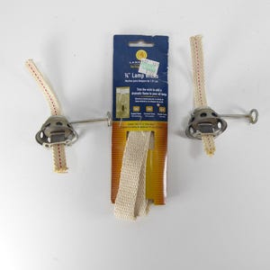 May include: Two metal oil lamp burners with off-white cotton wicks and a central retail package labeled 3/4 inch Lamp Wicks. The packaging is blue and orange with a yellow flame logo and includes a long, folded strip of replacement cotton wick.