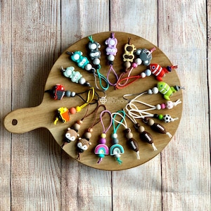 May include: A selection of colourful silicone phone charms featuring various animal and object designs. The charms are attached to a string and have wooden beads. The charms are displayed on a wooden chopping board.