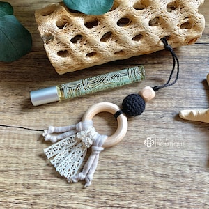 May include: A wooden ring with a black crocheted bead and a beige tassel hangs from a black cord. The ring is attached to a small bottle of essential oil with a green and white label.
