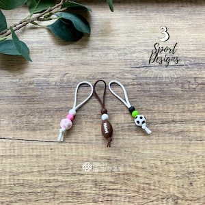 May include: Three zipper pulls with sports-themed charms. A pink soccer ball, a brown football, and a black and white soccer ball.