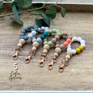 May include: Five colorful silicone and wooden bead keychains with gold-tone clasps. The keychains are arranged in a row on a wooden surface. The colors of the beads are teal, green, pink, orange, and yellow.