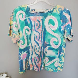 May include: A short-sleeve t-shirt featuring an abstract print in shades of teal, yellow, pink, and white. The design includes large swirling shapes, vertical wavy lines, and a small graphic of a vehicle. The shirt has a round neckline.