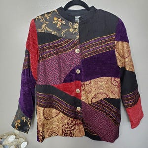 May include: A long-sleeve button-up jacket featuring a patchwork design of various fabrics in shades of black, deep purple, red, and gold. The garment includes panels with intricate floral and geometric patterns, a mandarin collar, and five round buttons.
