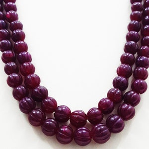 Puede incluir: Un collar de varias hebras con cuentas redondas y acanaladas de color burdeos intenso. Las cuentas están ensartadas en una disposición en capas, creando una textura consistente en toda la pieza. El color rojo oscuro es uniforme en todas las esferas.