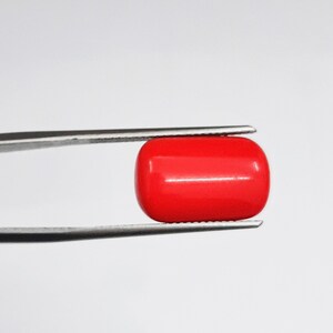 May include: A vibrant, polished red gemstone, rectangular with rounded edges, is held by silver tweezers against a white background. The gemstone's smooth surface reflects light, showcasing its rich color and shape. This item is likely used in jewelry.