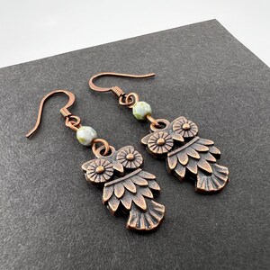 May include: A pair of copper owl earrings with green and white beads. The owls have detailed wings and eyes.