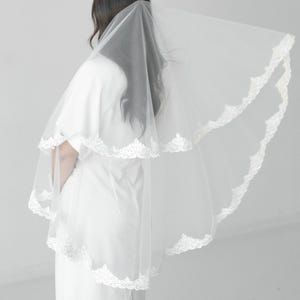 May include: A two-tier white bridal veil featuring a scalloped lace trim along the edges. The sheer tulle fabric is layered, with the shorter tier resting over the longer tier, creating a soft, flowing appearance against the white dress.