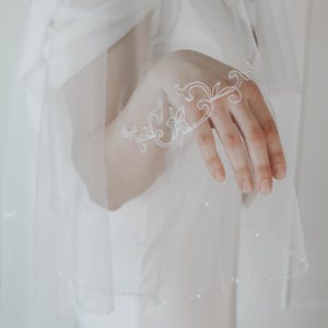 May include: A sheer white wedding veil with a delicate floral embroidery design on the edge. The veil is layered and has a subtle sparkle along the bottom edge. The embroidery is white and features a floral and swirl pattern.