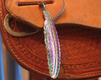 May include: A close-up of a leather saddle with a metal feather charm hanging from it. The feather is iridescent and has a purple, green, and blue color scheme.