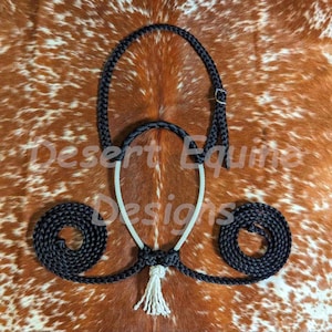 May include: A black and white rope horse halter with a matching lead rope, featuring a decorative tassel. The halter has a buckle closure and is displayed on a brown and white patterned surface. The text "Desert Equine Designs" is visible.