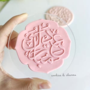 May include: A pink silicone stamp with Arabic calligraphy that reads "Eid Mubarak". The stamp is on a clear acrylic base and is being held by a hand.