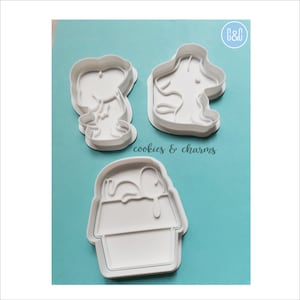 May include: Three white plastic cookie cutters shaped like a dog, a dog in a doghouse, and a dog sleeping in a doghouse. The cutters are on a light blue background with the text "cookies & charms" printed below the cutters.