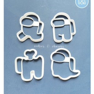 May include: Four white plastic cookie cutters shaped like characters from the video game Among Us. The cutters are on a blue background. The text "cookies & charms" is visible in the image.