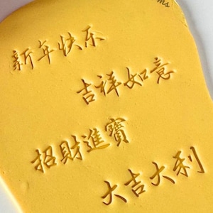 Könnte beinhalten: Ein gelber Teig in unregelmäßiger Form mit geprägten chinesischen Schriftzeichen. Der Text enthält Sätze wie „Frohes neues Jahr“ und „Viel Glück“. Die Worte „cookies & charms“ sind oben rechts aufgedruckt.