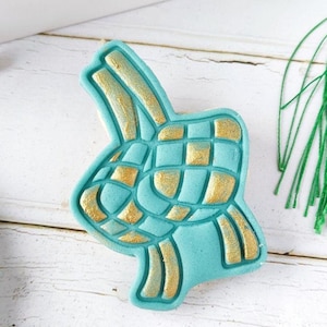 Peut inclure: Un biscuit turquoise et or décoré d'un motif tissé, ressemblant à un ketupat malaisien traditionnel, un plat de riz. Le biscuit est sur une surface en bois blanc. Le texte "A Baked Canvas" est visible en bas de l'image.