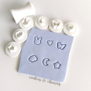 Puede incluir: Un juego de 10 cortadores de galletas de plástico blanco con varias formas, incluyendo una estrella, un corazón, una mariposa, un conejo, una nube, una luna y un círculo. Los cortadores están dispuestos en una estera de silicona azul claro con el texto "cookies & charms".