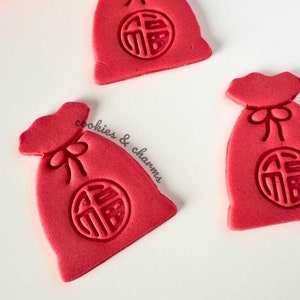 May include: Three red cookies shaped like money bags, each with a circular emblem and a bow at the top. The cookies are arranged on a white surface, with a few red flower petals in the background. The cookies are from "cookies & charms".