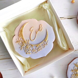May include: A white box with a clear lid contains a decorated cookie. The cookie is shaped like a cloud with a gold crescent moon and floral design. The word "Eid" is written in gold script. The text "A Baked Canvas" is visible.