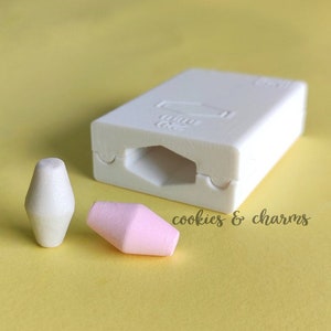 Puede incluir: Un molde de silicona blanco con una cavidad hexagonal, junto a dos pequeños amuletos geométricos en forma de lágrima, uno blanco y otro rosa. Los amuletos y el molde están sobre una superficie amarilla. El texto "cookies & charms" es visible.