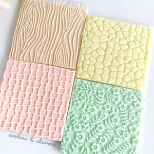 May include: Four square silicone baking mats with different embossed patterns: wood grain, stone, checkered, and floral. The mats are light brown, yellow, pink, and green. The text "cookies & charms" is printed on the bottom left corner.