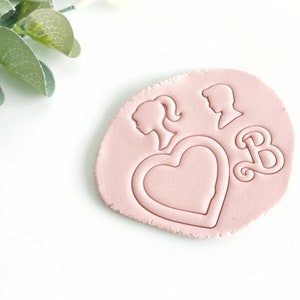Könnte beinhalten: Ein rosa Stempel aus Polymer Clay mit einem Herzen, zwei Profilsilhouetten und dem Buchstaben "B".