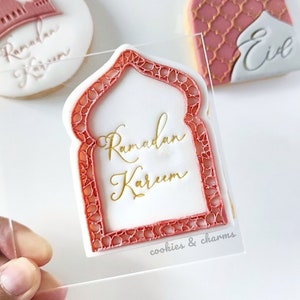 Può includere: Un biscotto bianco con un design ad arco rosso e bianco. Il biscotto è decorato con lettere dorate che recitano "Ramadan Kareem". Il biscotto è su una superficie acrilica trasparente con il testo "cookies & charms" stampato su di essa.