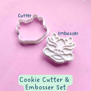May include: White plastic cookie cutter and embosser set. The cutter is shaped like a dragon's head. The embosser is shaped like a dragon's head with a detailed design. The set is on a pink background. Cookie Cutter & Embosser Set.