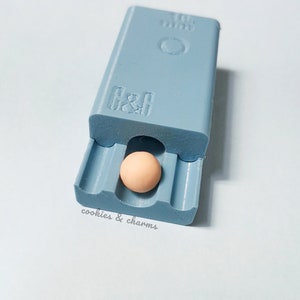 May include: A light blue plastic mold with the text "cookies & charms" printed on the bottom. A small, round, light brown bead is inside the mold.