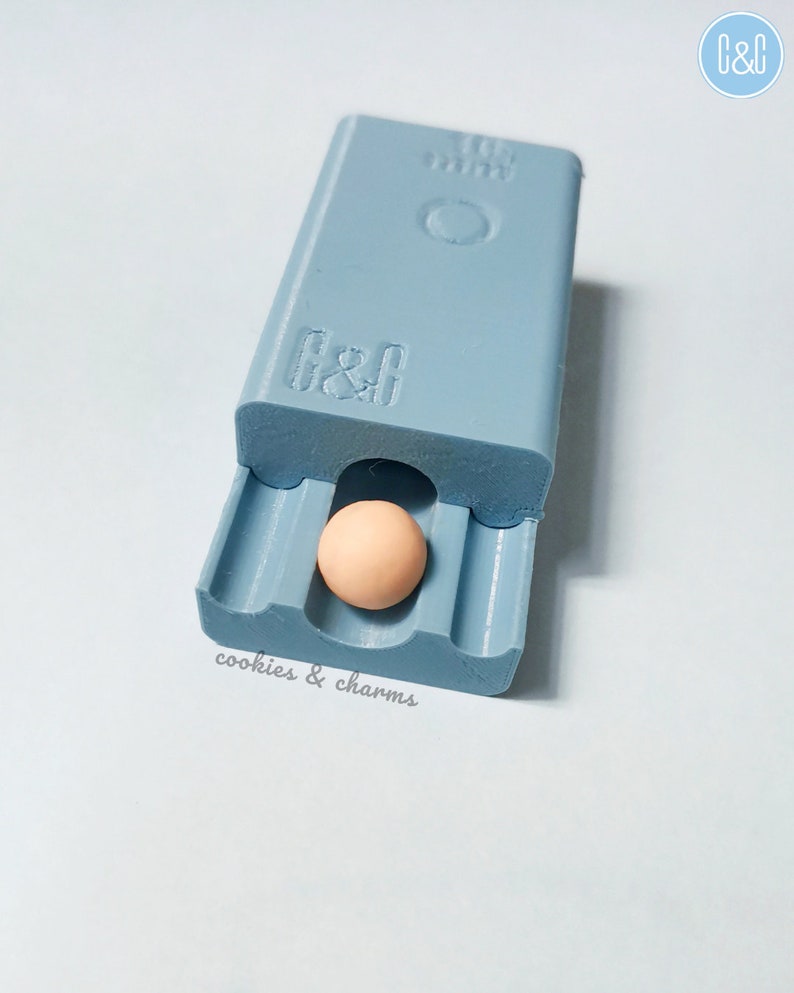 May include: A light blue plastic mold with the text "cookies & charms" printed on the bottom. A small, round, light brown bead is inside the mold.