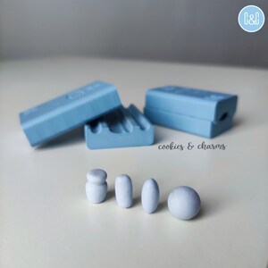 May include: Four light blue clay shapes, including a sphere, a cylinder, and two ovals, are arranged in a row on a white surface.  Two light blue rectangular molds are in the background.  The text "cookies & charms" is printed on the surface.