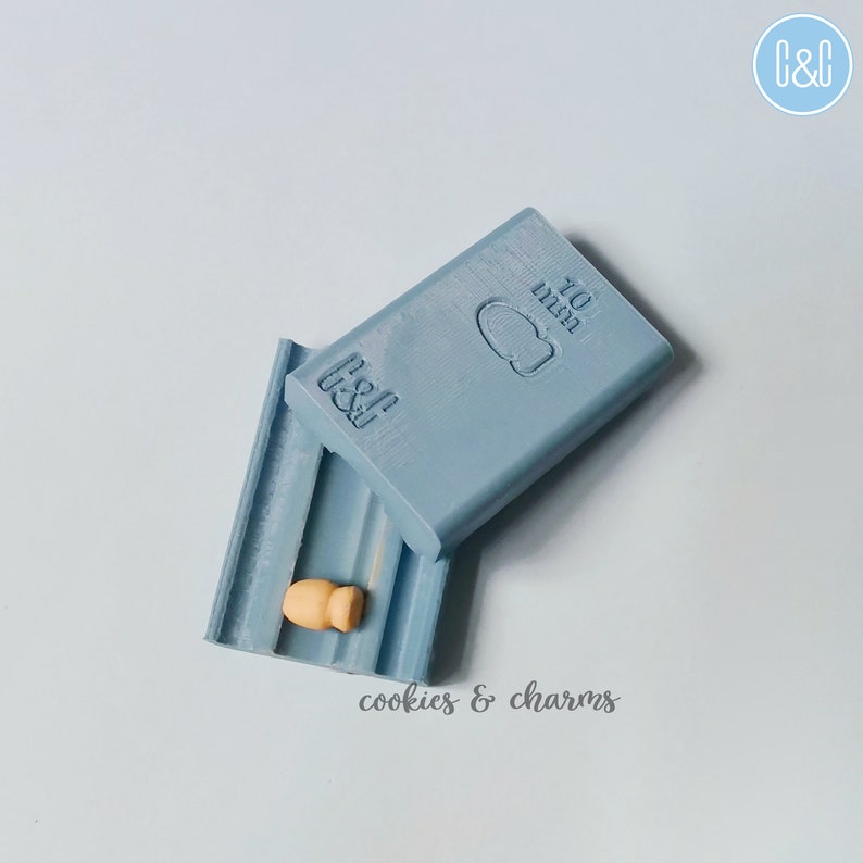 May include: A light blue plastic mold with the text "C&C" and "10 mm" on it. The mold is open and a small, light brown clay figurine is inside. The text "cookies & charms" is written below the mold.