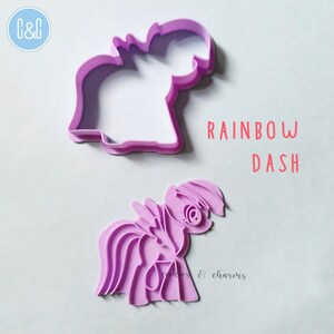 Puede incluir: Cortador de galletas de pl&aacute;stico morado con forma de Rainbow Dash de My Little Pony. El cortador est&aacute; en dos piezas, una para el contorno y otra para los detalles. El texto "RAINBOW DASH" est&aacute; impreso sobre un fondo blanco.