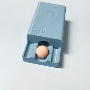 May include: A light blue plastic box with a small, round, light brown object inside. The box has the text "G&G" and "10 mm" printed on it.