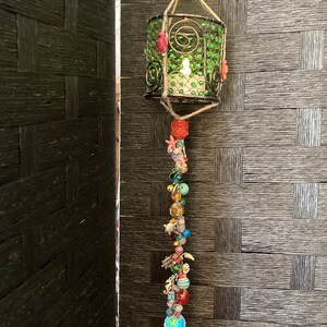 May include: A green beaded candle lantern with a long beaded chain hanging from it. The chain is made of various colored beads, including red, orange, yellow, green, blue, and purple. The lantern is hanging from a woven wall.